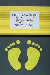 Photo of Card with phrase Big Journeys Begin With Small Steps and paper feet on dark background, top view. Motivational quote