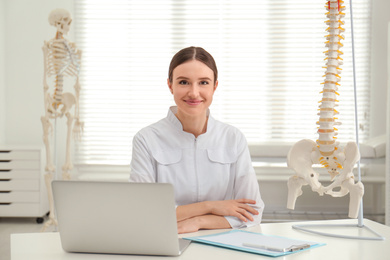 Female orthopedist with laptop near human spine model in office