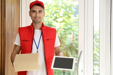 Happy young courier with parcel and tablet in doorway. Space for text