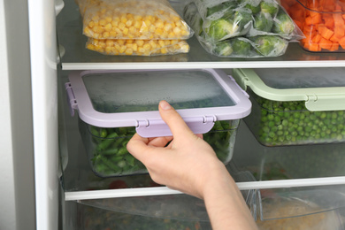 Woman putting container with green beans in refrigerator with frozen vegetables, closeup