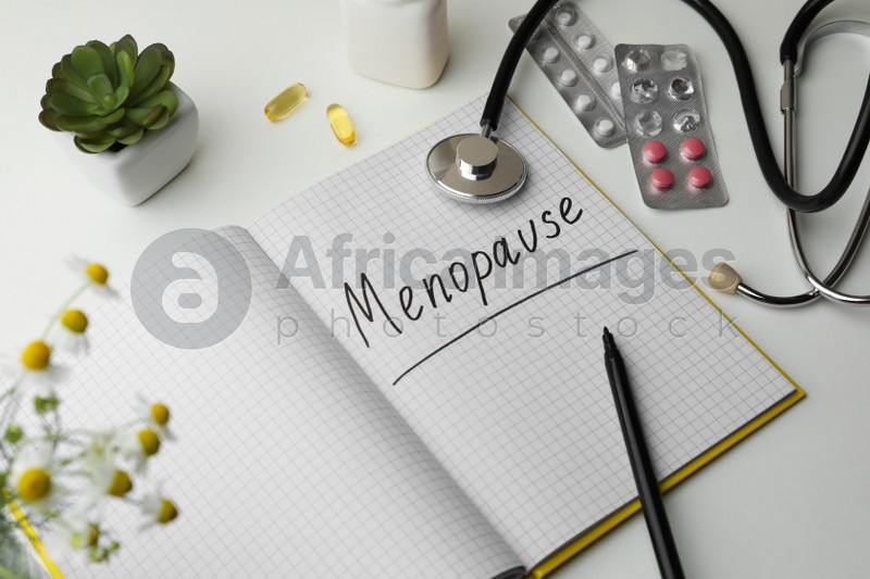 Photo of Notebook with word Menopause, stethoscope, pills and houseplant on white background, closeup