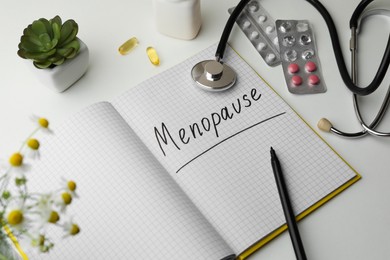 Notebook with word Menopause, stethoscope, pills and houseplant on white background, closeup