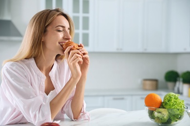Concept of choice between healthy and junk food. Woman eating croissant at white table in kitchen