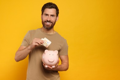 Happy man putting money into piggy bank on orange background, space for text