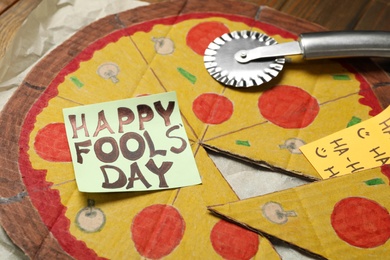 Cardboard pizza and Happy Fools' Day note on wooden table, closeup
