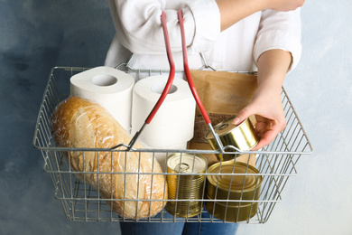 Woman holding shopping basket with products and toilet paper rolls on light blue background, closeup. Panic caused by virus