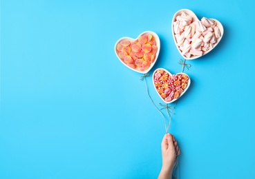 Woman holding twine near heart shaped bowls full of sweets imitating balloons on light blue background, top view. Space for text