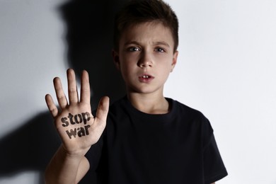 Unhappy little boy showing palm with phrase Stop War near white wall