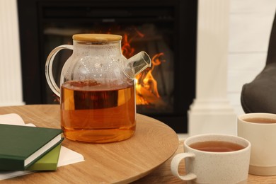Teapot with cups of hot drink and books on wooden tables near decorative fireplace in room. Interior design