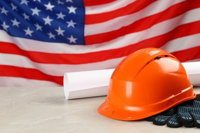 Photo of Orange protective hard hat, gloves, construction drawing on light grey table against American flag. Space for text