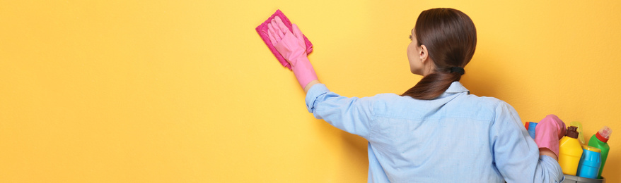 Woman with basin of detergents cleaning yellow wall, space for text. Banner design