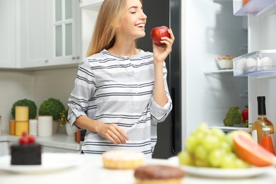 Concept of choice between healthy and junk food. Woman eating fresh apple near refrigerator in kitchen