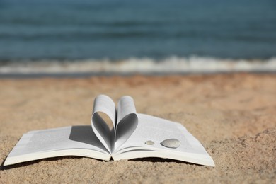 Open book with pages folded in heart shape on sandy beach near sea