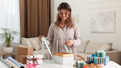 Beautiful young woman decorating gift box with ribbon at table in living room