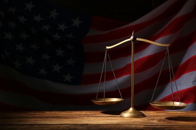 Scales of justice on wooden table against American flag in darkness, space for text