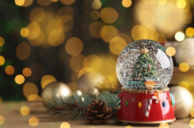 Beautiful snow globe and Christmas decor on wooden table against blurred festive lights, space for text. Bokeh effect