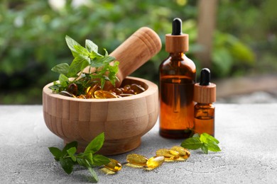 Wooden mortar with fresh green herbs, extracts and capsules on light grey table outdoors