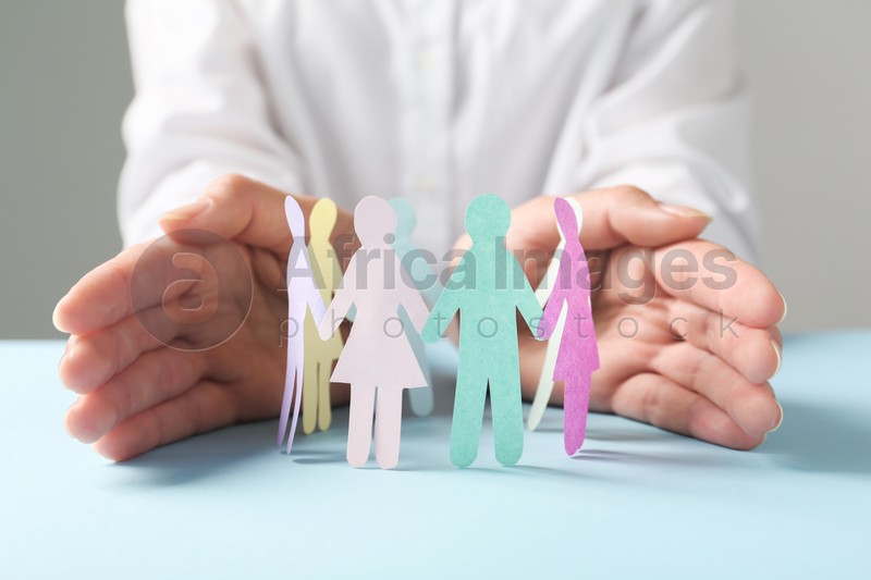 Photo of Woman protecting paper human figures on light blue table against grey background, closeup. Diversity and inclusion concept