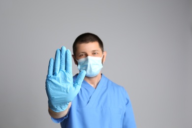 Doctor in protective mask showing stop gesture on grey background. Prevent spreading of COVID‑19