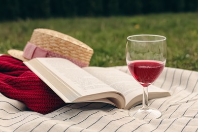 Blanket with glass of red wine, book, sweater and hat on green grass. Picnic season