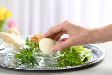 Woman holding traditional egg over Passover (Pesach) Seder plate on table, closeup