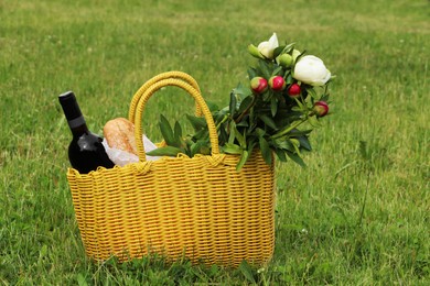 Yellow wicker bag with wine, bread and flowers on green grass outdoors. Picnic season