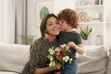 Photo of Little son kissing and congratulating his mom with Mother`s day at home. Woman holding bouquet of flowers