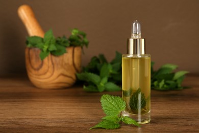 Glass bottle of nettle oil with dropper and leaves on wooden table against brown background, space for text