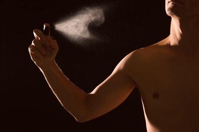 Young man spraying perfume on black background, closeup