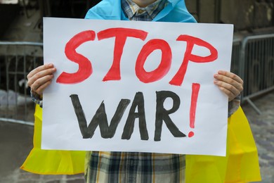 Woman wrapped in Ukrainian flag holding poster with words Stop War outdoors, closeup