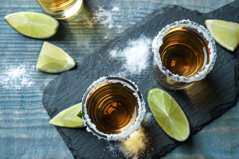 Mexican Tequila shots, lime slices and salt on blue wooden table, flat lay