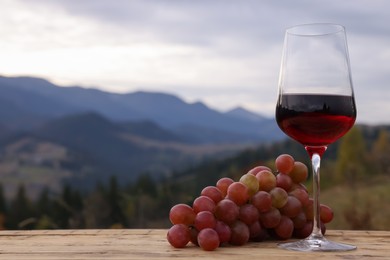 Photo of Glass of tasty red wine and grapes on wooden table against mountain landscape. Space for text