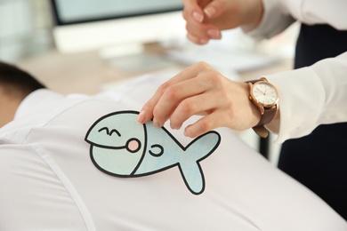 Young woman sticking paper fish to colleague's back in office, closeup. Funny joke