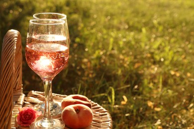 Photo of Glasses of delicious rose wine, flower and peaches on wicker basket outside. Space for text