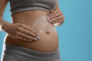 Pregnant woman applying cosmetic product on belly against light blue background, closeup. Space for text