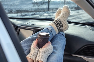 Young woman in warm socks holding her legs on car dashboard and drinking coffee. Cozy atmosphere