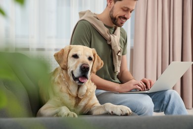 Photo of Man using laptop on sofa near his cute Labrador Retriever at home