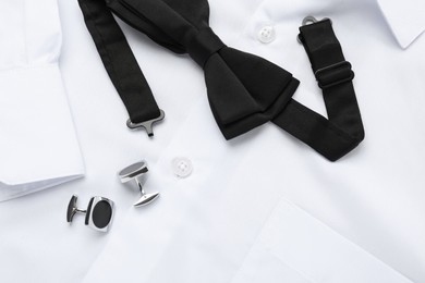 Stylish black bow tie and cufflinks on white shirt, top view