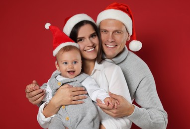 Happy couple with cute baby wearing Santa hats on red background. Christmas season