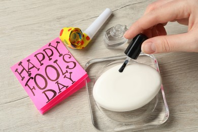 Woman applying transparent nail polish onto soap bar near paper with words Happy Fool's Day at wooden table, closeup