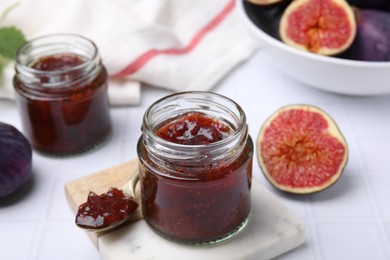 Photo of Glass jars of tasty sweet fig jam and fruits on white tiled table
