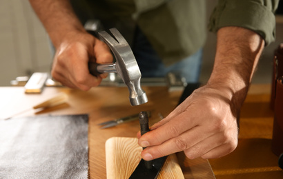 Photo of Man making holes in leather belt with punch and hammer at workshop, closeup