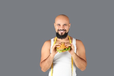 Overweight man with hamburger and measuring tape on gray background
