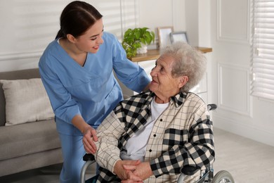 Young caregiver assisting senior woman in wheelchair indoors. Home health care service