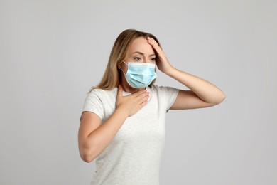 Young woman in medical mask suffering from pain during breathing on light grey background
