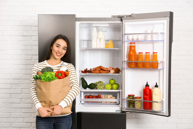 Young woman with paper bag full of products near open refrigerator indoors