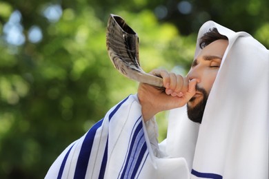 Jewish man in tallit blowing shofar outdoors. Rosh Hashanah celebration