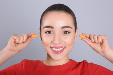 Young woman inserting foam ear plugs on grey background