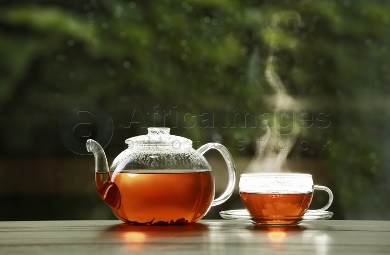 Teapot and cup of hot tea on wooden table against blurred background, space for text