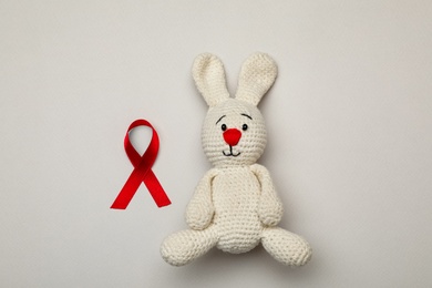Cute knitted toy bunny and red ribbon on beige background, flat lay. AIDS disease awareness